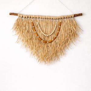 K&ouml;nnte beinhalten: Wandbehang aus Naturfasern in Dreiecksform. Er hat einen Holzast an der Oberseite, einen gewebten Abschnitt und eine Kaskade aus getrocknetem, gras&auml;hnlichem Material. Dekorative Holzperlen und wei&szlig;e Perlen sind in der Mitte aufgeh&auml;ngt.