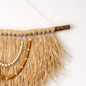K&ouml;nnte beinhalten: Wandbehang aus Naturfasern mit Holzstab und Schnur zur Aufh&auml;ngung. Das St&uuml;ck hat ein gewebtes Design mit langen, ausgefransten Str&auml;hnen in Beige- und Braunt&ouml;nen, akzentuiert mit Holzperlen.