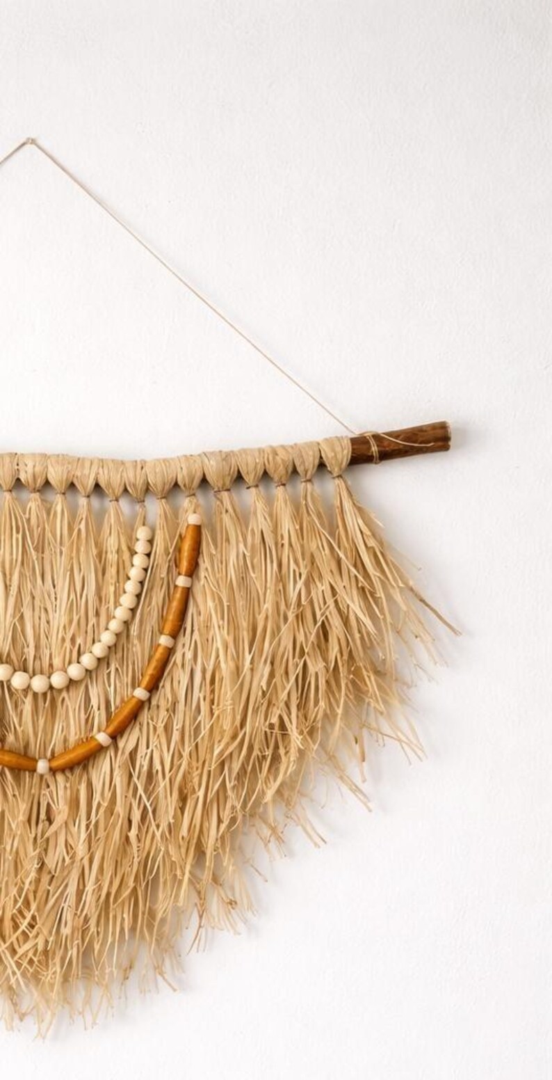 K&ouml;nnte beinhalten: Wandbehang aus Naturfasern mit Holzstab und Schnur zur Aufh&auml;ngung. Das St&uuml;ck hat ein gewebtes Design mit langen, ausgefransten Str&auml;hnen in Beige- und Braunt&ouml;nen, akzentuiert mit Holzperlen.