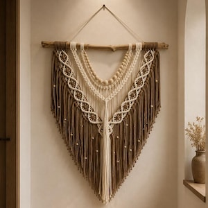 Puede incluir: Un tapiz de macramé en tonos marrón y crema, suspendido de una rama de madera natural. El diseño presenta intrincados patrones de nudos, detalles de cuentas y un fondo con flecos, creando una estética bohemia.