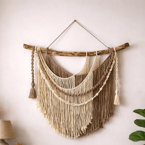 May include: A macrame wall hanging with a natural wooden branch and beige and brown cotton cord. The design includes layered fringe and knotted details, creating a bohemian aesthetic. The measurements are in centimetres.