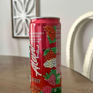 Puede incluir: Una lata de bebida energ&eacute;tica roja decorada con brillantes pedrer&iacute;a. La lata presenta el texto "Strawberry Sunrise" y "Energy". La pedrer&iacute;a forma dise&ntilde;os de fresas y hojas. La parte inferior de la lata es azul claro.