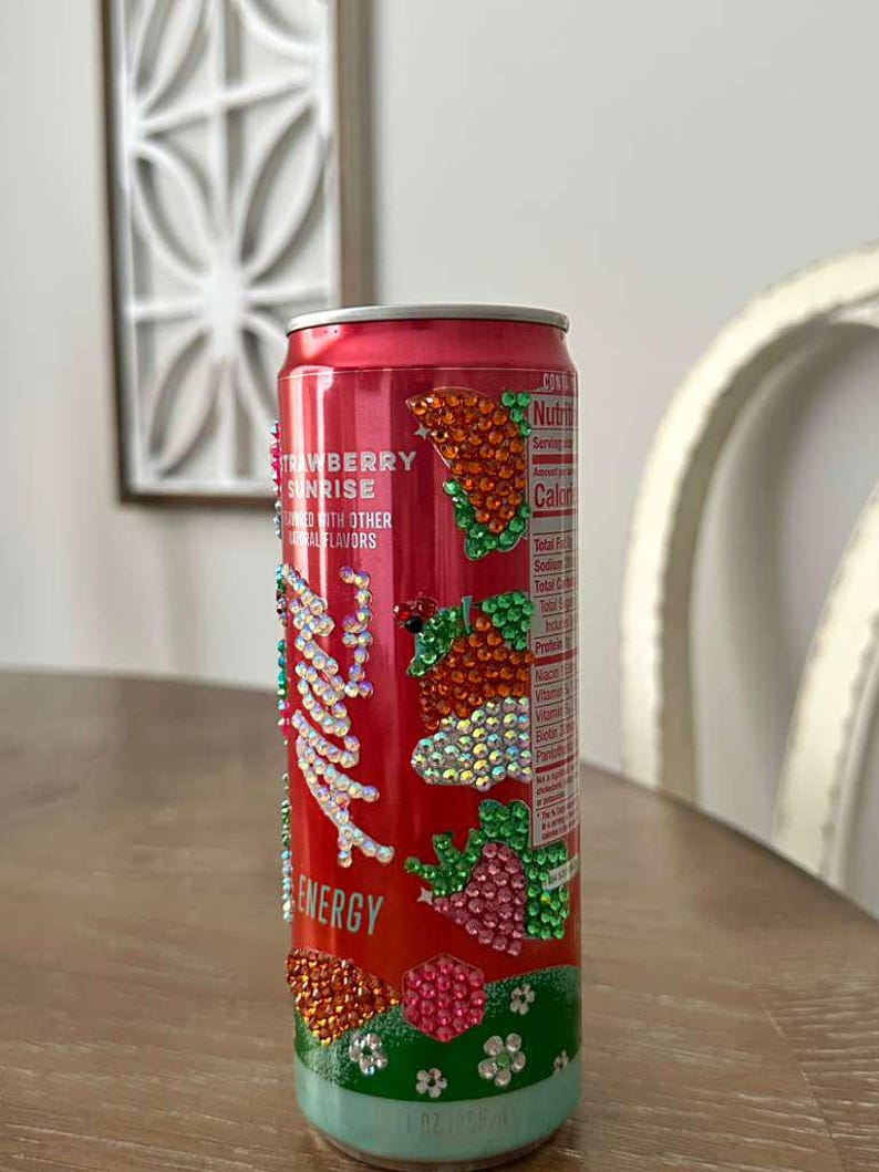 Puede incluir: Una lata de bebida energ&eacute;tica roja decorada con brillantes pedrer&iacute;a. La lata presenta el texto "Strawberry Sunrise" y "Energy". La pedrer&iacute;a forma dise&ntilde;os de fresas y hojas. La parte inferior de la lata es azul claro.