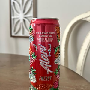Puede incluir: Una lata de bebida energ&eacute;tica roja con el texto "Strawberry Sunrise" y "Alani" en cursiva. La lata est&aacute; decorada con pedrer&iacute;a colorida y una base azul claro.