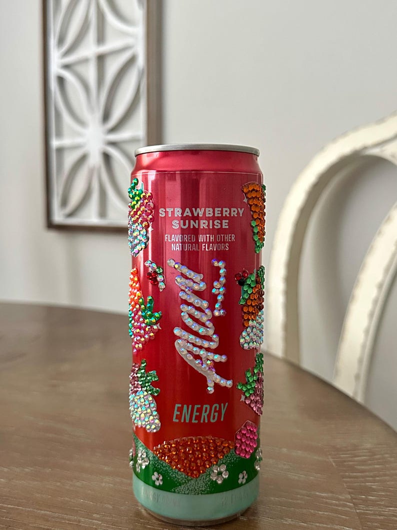 Puede incluir: Una lata de bebida energ&eacute;tica roja con el texto "Strawberry Sunrise" y "Alani" en cursiva. La lata est&aacute; decorada con pedrer&iacute;a colorida y una base azul claro.