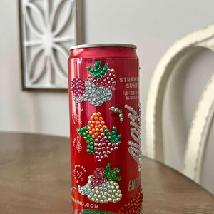 Puede incluir: Una lata de bebida roja decorada con brillantes pedrer&iacute;a en un dise&ntilde;o de fresa. La lata presenta el texto "Strawberry Sunrise" y "Energy". La parte inferior de la lata es azul claro con detalles florales.
