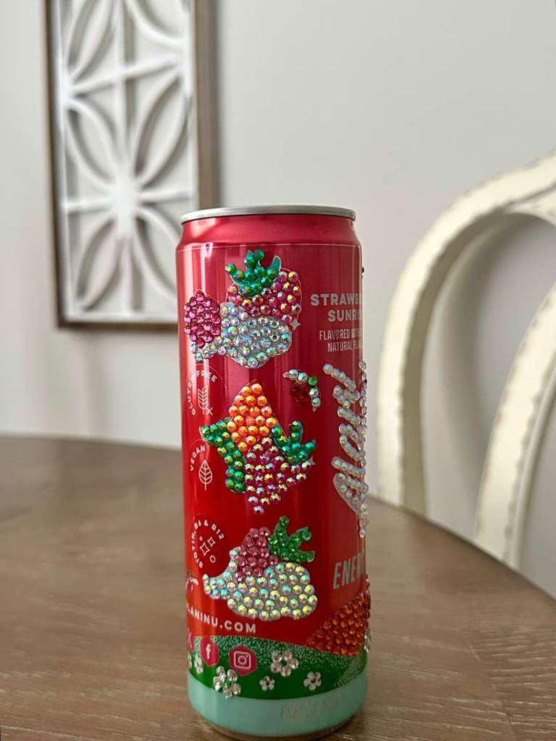 Puede incluir: Una lata de bebida roja decorada con brillantes pedrer&iacute;a en un dise&ntilde;o de fresa. La lata presenta el texto "Strawberry Sunrise" y "Energy". La parte inferior de la lata es azul claro con detalles florales.