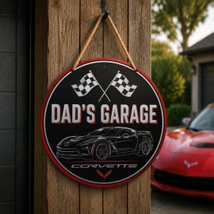 May include: A round black sign with a red border hanging by a rope. It features two crossed checkered flags, the text "DAD'S GARAGE" in white, a white line drawing of a Corvette sports car, the word "CORVETTE", and a small red emblem at the bottom.