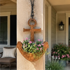 May include: A copper-colored metal anchor-shaped wall planter hanging from a black chain. The planter contains a cluster of small pink flowers and green foliage, filling the curved base of the anchor structure against a wooden post.