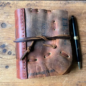 May include: A handmade leather journal with a baseball glove design. The journal is brown with a leather tie closure. A black pen with gold accents rests beside the journal. The journal has the words "ROMO" and "Professional Players" printed on it.