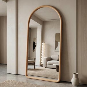May include: A full-length, arched-top mirror with a light wood frame. The mirror reflects a neutral-toned living space with a sofa, chair, and artwork. The mirror is leaning against a light-colored wall.