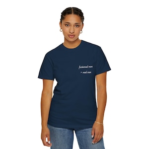 Puede incluir: Camiseta azul marino con el texto blanco "fictional men > real men". La camiseta, de cuello redondo, se lleva con vaqueros azules. La persona lleva una pulsera dorada.