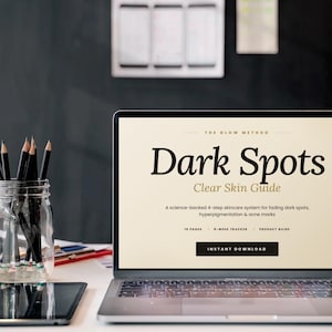 May include: A laptop screen shows a skincare guide titled "Dark Spots". The text describes a 4-step system for fading dark spots and acne marks. A jar of pencils and a tablet are on the desk. The background is a dark wall.
