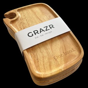 Könnte beinhalten: Ein Stapel hellfarbener Holzserviertabletts in abgerundeter rechteckiger Form mit einer kreisförmigen Aussparung für ein Glas. Ein weißes Papierband umschließt die Mitte mit dem schwarzen Text GRAZR Eat · Sip · Mingle und dem eingravierten One hand.