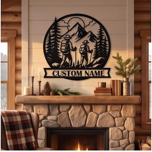 May include: A circular black metal wall art piece featuring a silhouette of two hikers with backpacks and trekking poles in front of mountains, pine trees, and a sun. Below the circular frame is a banner with the text CUSTOM NAME.