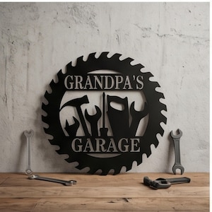 Könnte beinhalten: Ein kreisförmiges Wand-Schild aus schwarzem Metall in Form eines Sägeblatts mit dem Schriftzug "GRANDPA'S GARAGE" in der Mitte. Das Design zeigt Silhouetten von Hammer, Schraubendreher, Handsäge und zwei Schraubenschlüsseln zwischen den Wörtern.