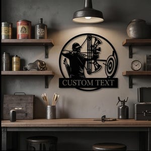 Peut inclure: Une décoration murale circulaire en métal noir représentant la silhouette d'un archer visant une cible avec un arc à poulies. Le design inclut un carquois avec des flèches au dos et une bannière rectangulaire avec le texte CUSTOM TEXT.