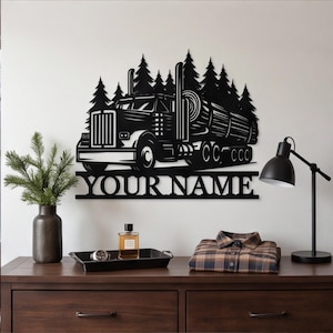 Op de afbeelding: Een zwart metalen wanddecoratie met een vrachtwagen die boomstammen vervoert voor een dennenbos. Onder de vrachtwagen bevindt zich een horizontale balk met de tekst YOUR NAME in een vetgedrukt schreeflettertype, als een gepersonaliseerd bord.