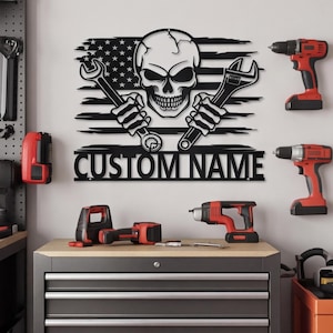 May include: A black metal wall sign featuring a stylized skull centered over a distressed American flag pattern. The skull holds two wrenches in its skeletal hands. Below the graphic, the text CUSTOM NAME is cut out in bold, block lettering.