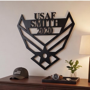 Peut inclure: Une enseigne murale en métal noir présentant le design de l'emblème de l'US Air Force avec le texte USAF, SMITH et 2020 découpé dans une police sans empattement. La structure géométrique et angulaire forme une silhouette d'aigle stylisée.