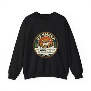 Op de afbeelding: Zwarte sweatshirt met een cirkelvormige afbeelding van een schaap, bergen en de tekst "99 SHEEP and GOD came back for the one that's me" en "GOD LOVE". De afbeelding is in bruin, groen en oranje.