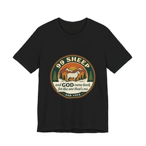 Op de afbeelding: Zwart T-shirt met een cirkelvormige afbeelding. De afbeelding bevat de tekst "99 Sheep", een schapenillustratie en de zin "and GOD came back for the one that's me". Het ontwerp bevat ook een berg en een zon, met "GOD LOVE" onderaan.