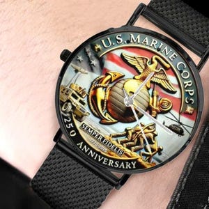 Op de afbeelding: Zwarte polshorloge met een ronde wijzerplaat met het embleem van het U.S. Marine Corps, een adelaar, wereldbol en anker, met de tekst "U.S. MARINE CORPS" en "250 ANNIVERSARY". Het horloge heeft een zwarte mesh band.