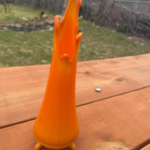 May include: An orange glass vase with a flared top and three small feet. The vase has a smooth, glossy finish and a vibrant, warm color. The vase is a decorative item, suitable for displaying flowers or as a standalone art piece.