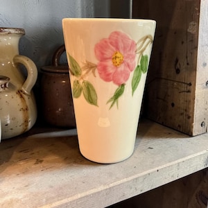 May include: A cream-colored ceramic vase with a pink rose and green leaves painted on the side. The vase has a slightly flared shape and is displayed on a wooden shelf.