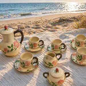 May include: A cream-colored tea set with pink rose floral designs. The set includes a teapot, sugar bowl, creamer, and six teacups with saucers. The teacups and teapot have dark brown handles. The set is arranged on a white blanket on a beach.