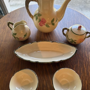 May include: A ceramic tea set featuring a cream-colored teapot with a lid, a creamer, a lidded sugar bowl, an elongated scalloped serving dish, and two small bowls. Each piece is decorated with hand-painted pink rose blossoms and green leaves.