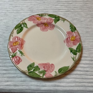 May include: A round, off-white ceramic plate with a floral design. Pink flowers with yellow centers and green leaves are arranged around the rim. The plate has a gold-colored edge.