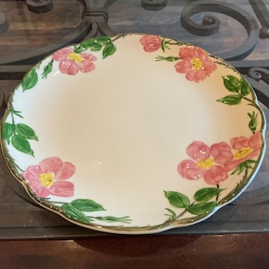 May include: A white ceramic plate with a scalloped edge, adorned with pink floral and green leaf designs. The plate has a gold trim and is likely used for serving food. The floral pattern is evenly spaced around the plate's edge.
