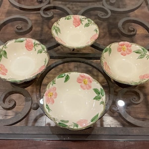 May include: Four off-white ceramic bowls with a scalloped edge. Each bowl features a pink floral design with green leaves. The bowls are arranged on a dark metal and glass table.