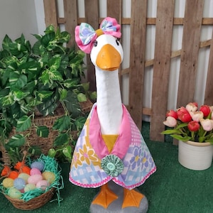 May include: A decorative goose statue with a white body, orange beak and feet, and a pink bow. The goose wears a pink, yellow, and blue patterned cape with a green button. The statue is standing on a gray stone base.