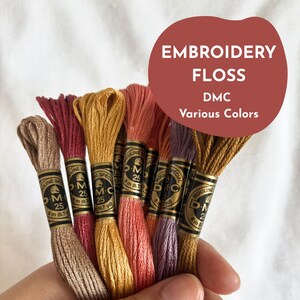 Può includere: Una mano tiene in mano diversi rocchetti di filo da ricamo DMC in vari colori. I rocchetti sono etichettati con il nome del marchio e il testo "EMBROIDERY FLOSS DMC Various Colors" è in un cerchio rosso.