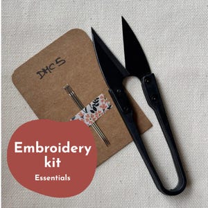 Può includere: Un kit da ricamo con forbici per filo nere, aghi e un cartoncino marrone. Il cartoncino presenta la scritta a mano "DHCS" ed è nastrato con nastro adesivo a motivi floreali. Un cerchio rosso con le parole "Embroidery kit Essentials" è in primo piano.