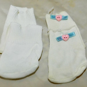 Puede incluir: Dos pares de calcetines blancos. Un par es liso con puño acanalado. El otro par presenta un adorno de cinta azul claro en el tobillo, acentuado con un pequeño botón rosa en forma de corazón centrado en cada cinta. Hechos de tela.