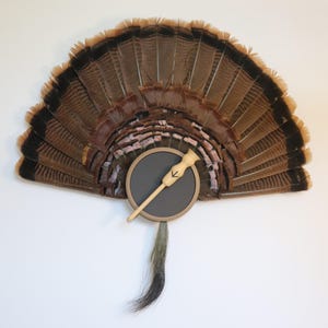 May include: A decorative fan made of brown turkey feathers, with a black and tan trim. A circular, dark gray element with a wooden handle and arrow design is attached to the fan. A tuft of gray and brown feathers hangs below.