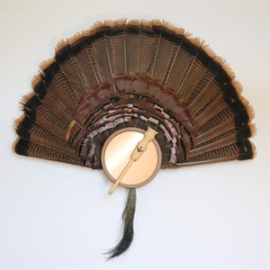 May include: A decorative fan made of turkey feathers, featuring a circular copper-colored element with a wooden handle. The feathers are primarily brown with black tips, and a dark tail hangs below. The fan is likely used for hunting or as a decorative item.