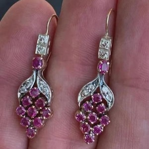 Peut inclure: Une paire de boucles d'oreilles pendantes composées d'un groupe de rubis ronds rose foncé disposés en grappe, agrémentées de petits diamants blancs transparents sertis sur un métal argenté, suspendues par une attache en crochet.
