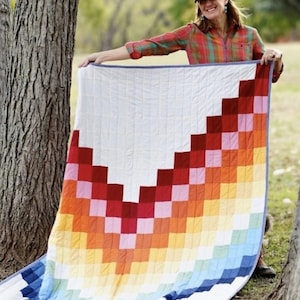 May include: A colorful quilt with a white top and a vibrant, pixelated rainbow design in shades of red, orange, yellow, and blue. The quilt is held up outdoors, showcasing its handmade quality and cheerful aesthetic.