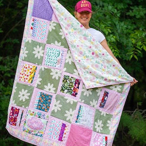 May include: A patchwork quilt featuring a variety of fabric squares in shades of green, pink, and white with floral and geometric patterns. One square includes the text Happy Birthday. The quilt has a light pink border and a floral backing.