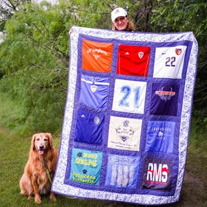May include: A rectangular memory quilt constructed from various athletic jerseys and graphic t-shirts in orange, red, white, blue, and black. The quilt features a blue and white patterned border and includes various printed logos, numbers, and text.