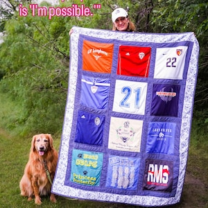 May include: A memory quilt constructed from various athletic jerseys and t-shirts in orange, red, white, blue, and black, featuring the text "You say impossible, but all I hear is 'I'm possible.'" The quilt has a blue and white patterned border.