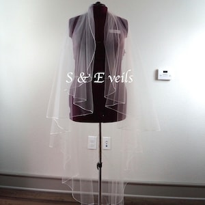 May include: A white single-tier wedding veil with a scalloped edge, displayed on a mannequin. The veil is made of soft tulle and has a delicate, flowing design. The text "S & E Veils" is visible on the mannequin's chest.