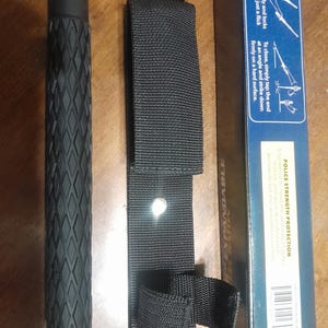 Może przedstawiać: Czarny, rozkładany pałka z teksturowanym uchwytem, czarną nylonową szlufką na pasek i pudełkiem produktu. Pudełko zawiera instrukcje i tekst "POLICE STRENGTH PROTECTION".