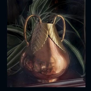 May include: A copper vase with a rounded body and two handles. The upper portion of the vase features two large, textured brass leaf-shaped overlays that extend from the rim down to the shoulder, providing a decorative contrast to the smooth copper.