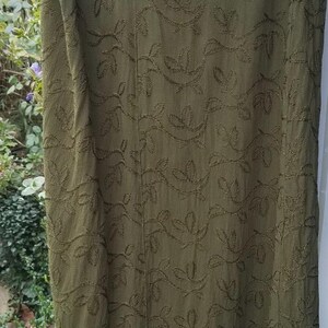 May include: Olive green skirt with embroidered leaf pattern. The skirt has a velvet hem. The skirt is hanging on a wooden hanger. The skirt is made of a soft, flowing fabric.