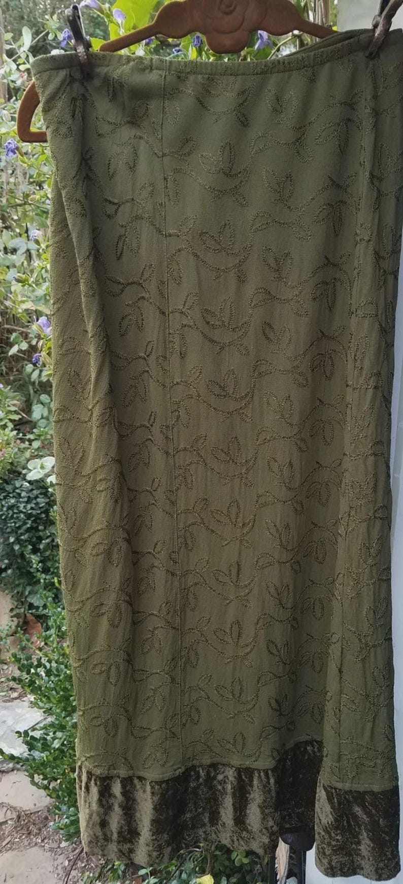 May include: Olive green skirt with embroidered leaf pattern. The skirt has a velvet hem. The skirt is hanging on a wooden hanger. The skirt is made of a soft, flowing fabric.